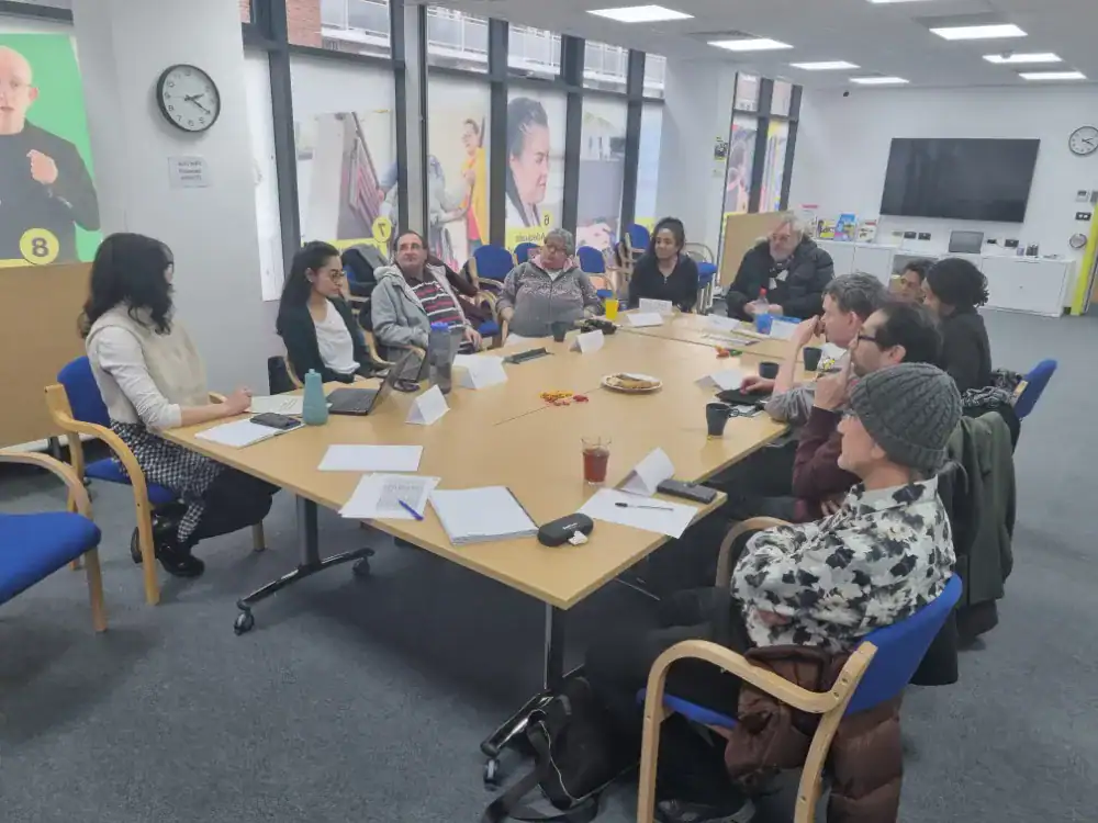 A group of people sit around a rectangular table in a meeting room, engaged in discussion. Papers, drinks, and snacks are on the table. Large windows and posters are visible in the background.