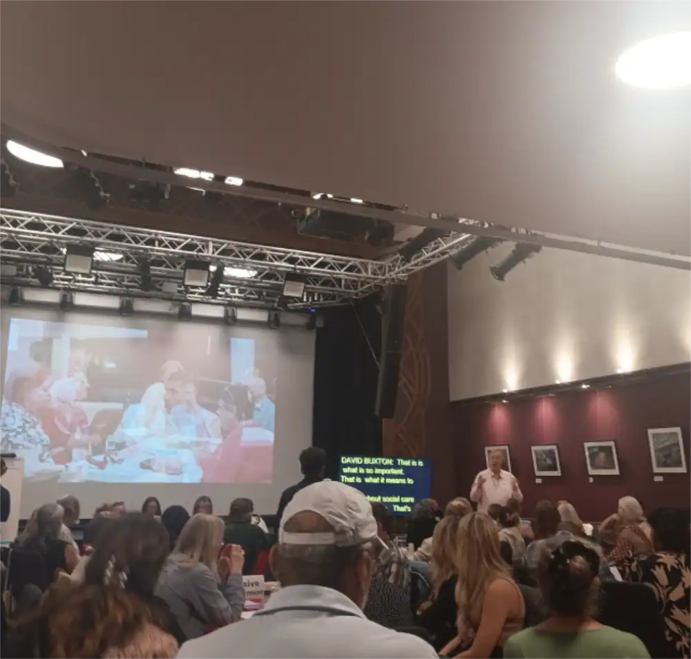 A speaker presents to an audience in a conference room; a projected image and live captions are visible behind him, with attendees seated at tables in the foreground.
