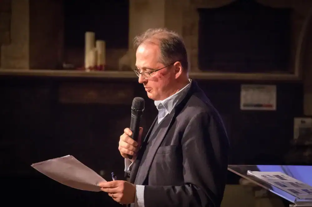 A man in a suit holding a microphone reads from a sheet of paper while standing indoors, with candles and papers visible in the background.
