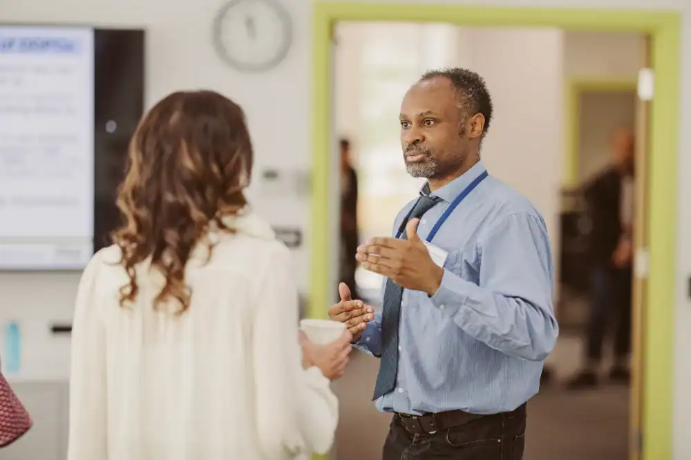 A man in a blue shirt and tie speaks to a woman holding a cup in an office setting, with a clock and a digital display visible in the background.