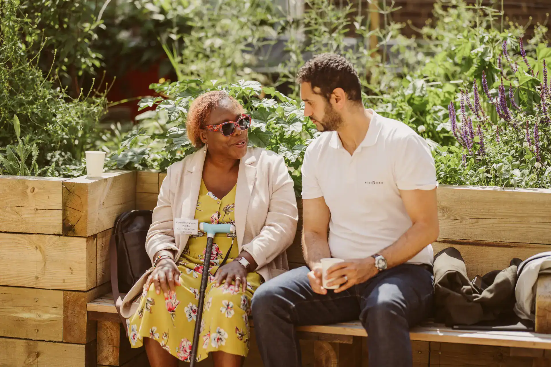 Two people sit on a wooden bench outdoors, engaged in conversation, with greenery in the background. Both hold cups and appear relaxed.