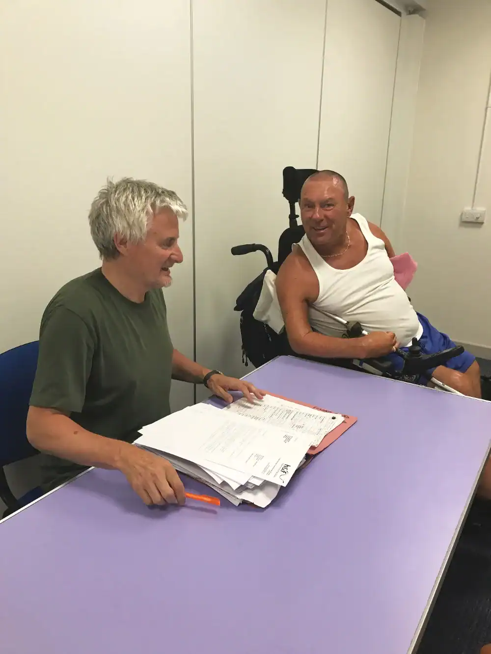 Two men sit at a purple table; one is holding paperwork and a pen, while the other, in a wheelchair, looks on and smiles.