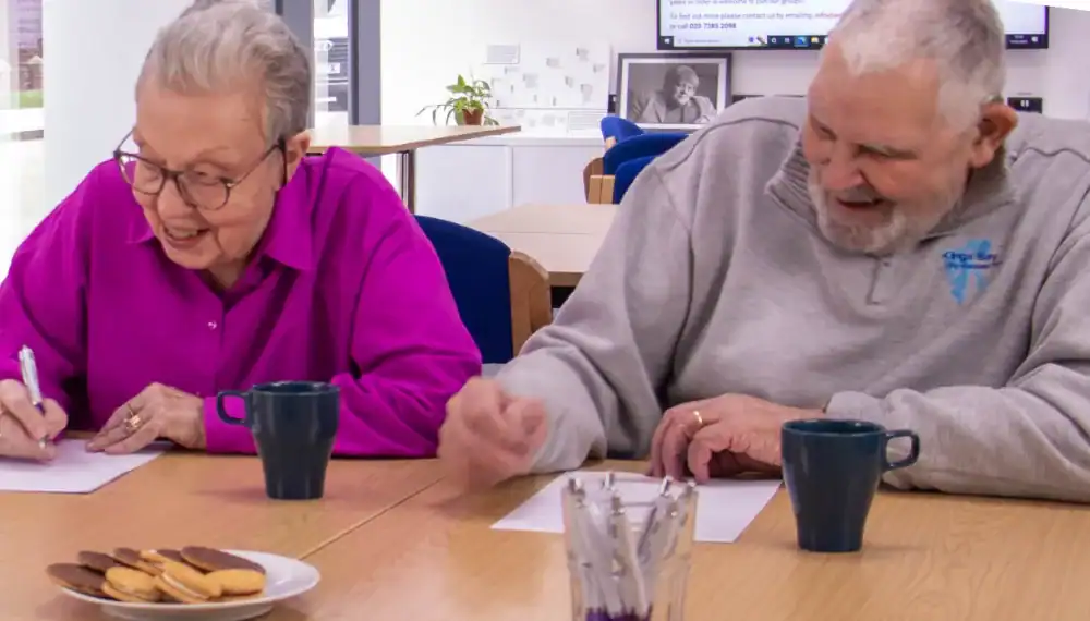 Two older adults sit at a table with mugs and cookies, writing on paper and smiling in a bright indoor setting.